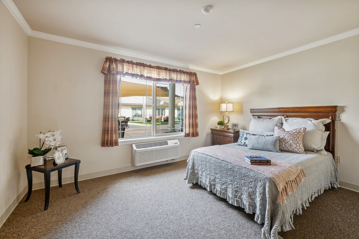 A cozy bedroom with a neatly made bed, featuring a large window with patterned curtains and warm lighting from a lamp on the bedside table.