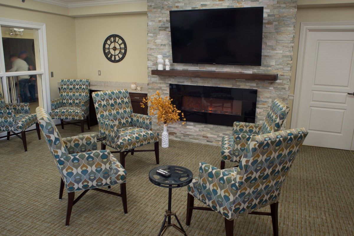 A cozy living room with patterned armchairs arranged around a modern fireplace and a large television above.