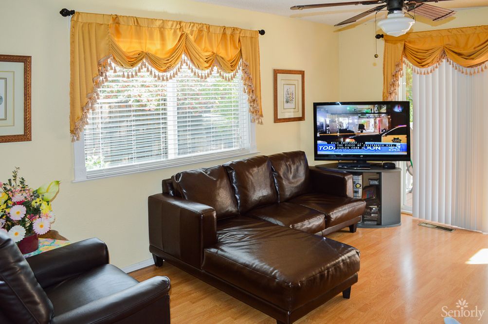 A cozy living room with a brown leather sofa, a TV, and large windows with yellow curtains.