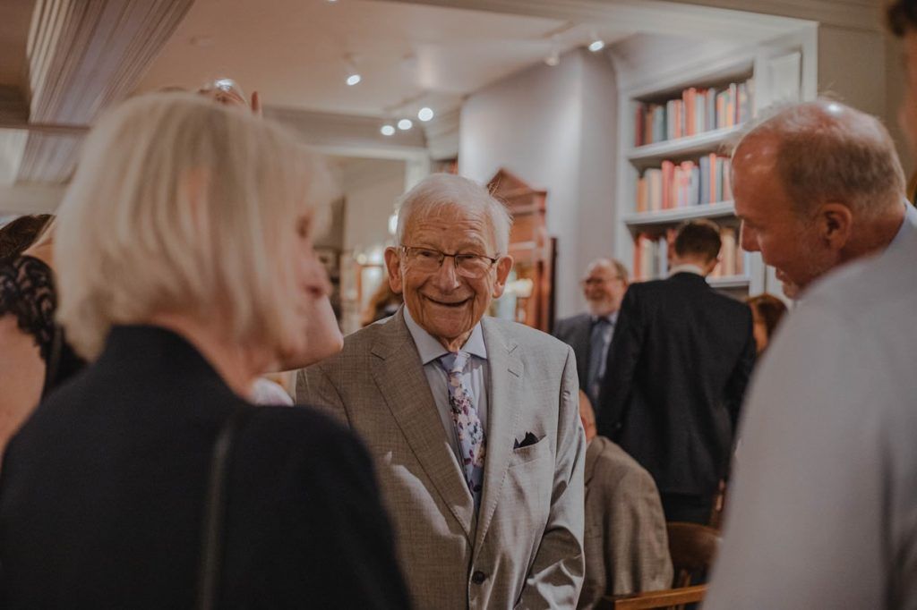 An elderly man in a suit engages in conversation at an event surrounded by books.