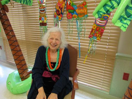 An elderly woman smiles while seated in a vibrant room decorated with colorful items.