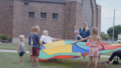 A joyful gathering with children and an adult playing with a colorful parachute in an outdoor setting.