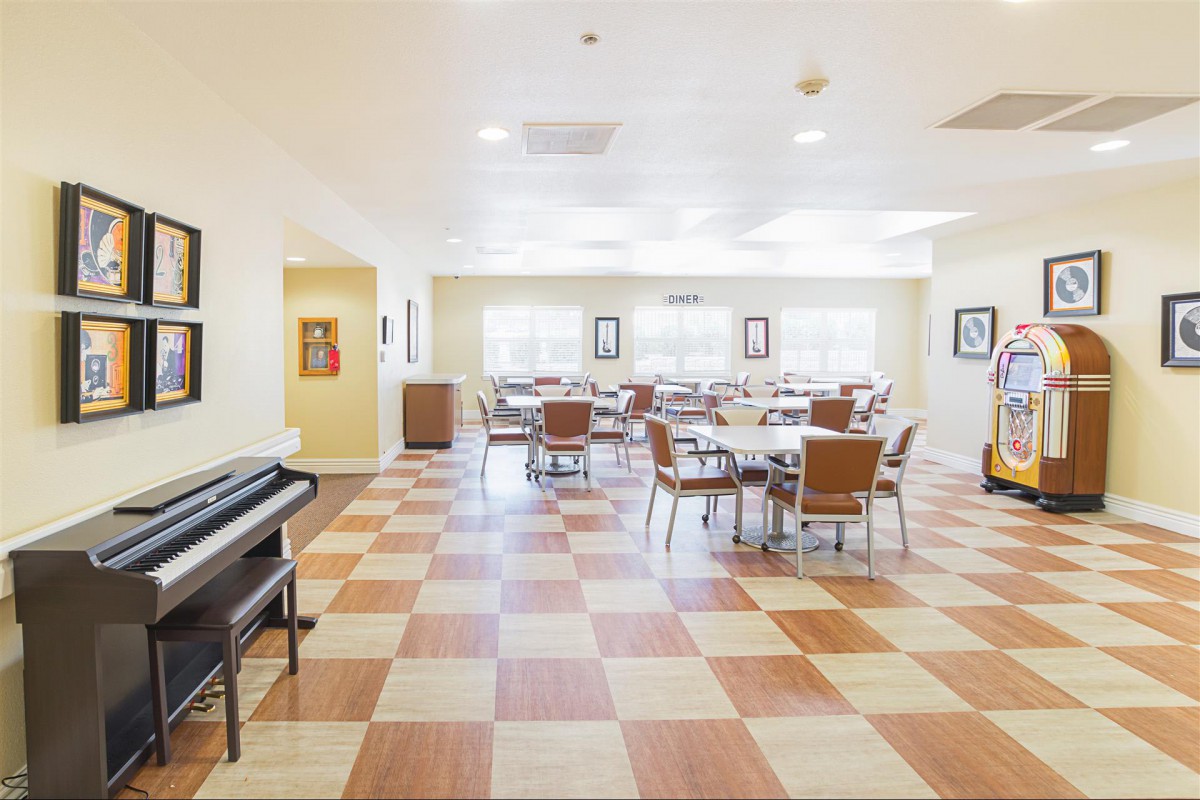 A well-lit dining area with checkered flooring, featuring multiple tables and chairs, and a piano in the corner.