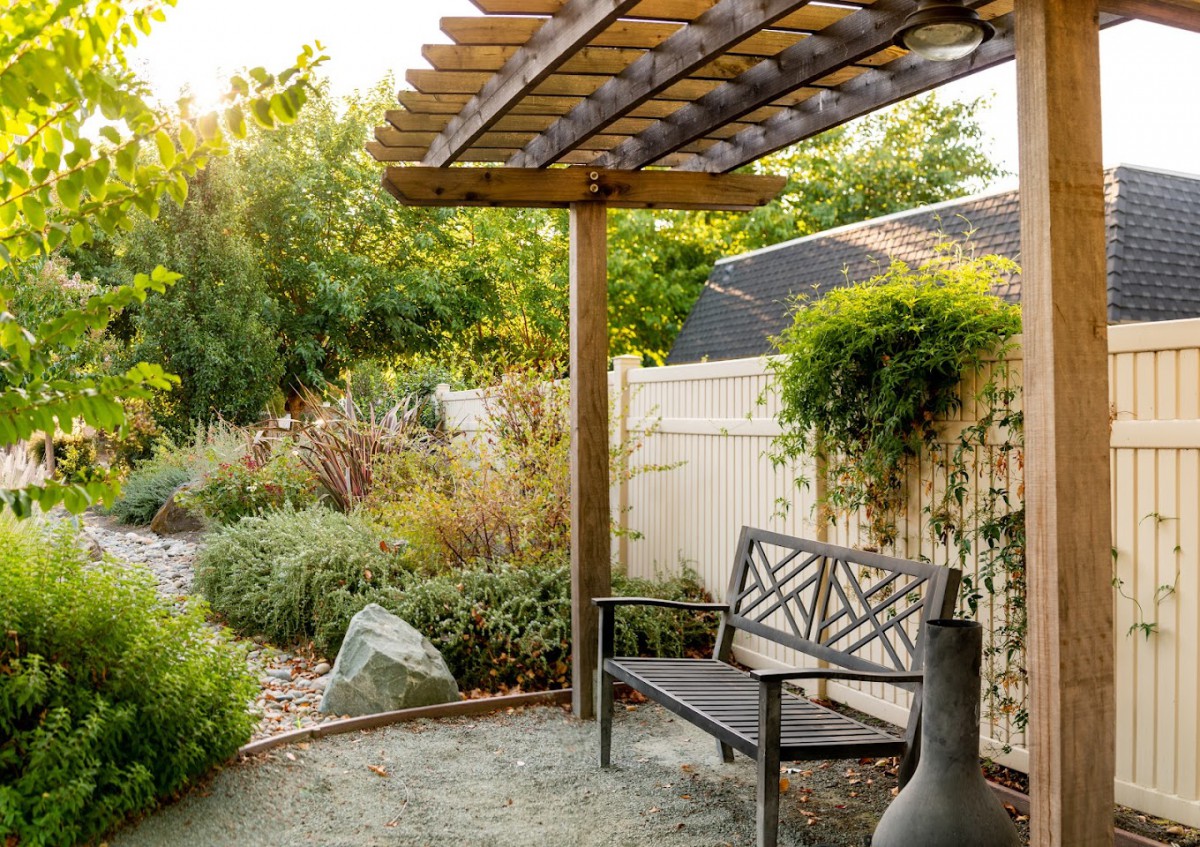 A serene garden setting featuring a wooden pergola with a decorative bench underneath, surrounded by lush greenery.