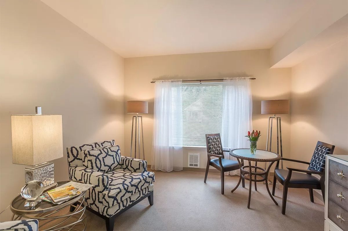 A cozy living room with modern furnishings, including an armchair, a small round table with chairs, and large windows adorned with curtains.