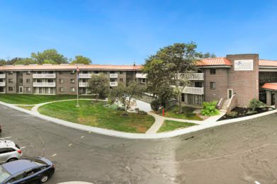 A multi-story apartment complex with green lawns and trees under a clear blue sky.