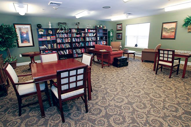 A cozy library reading room with bookshelves, tables, and chairs on a carpeted floor.