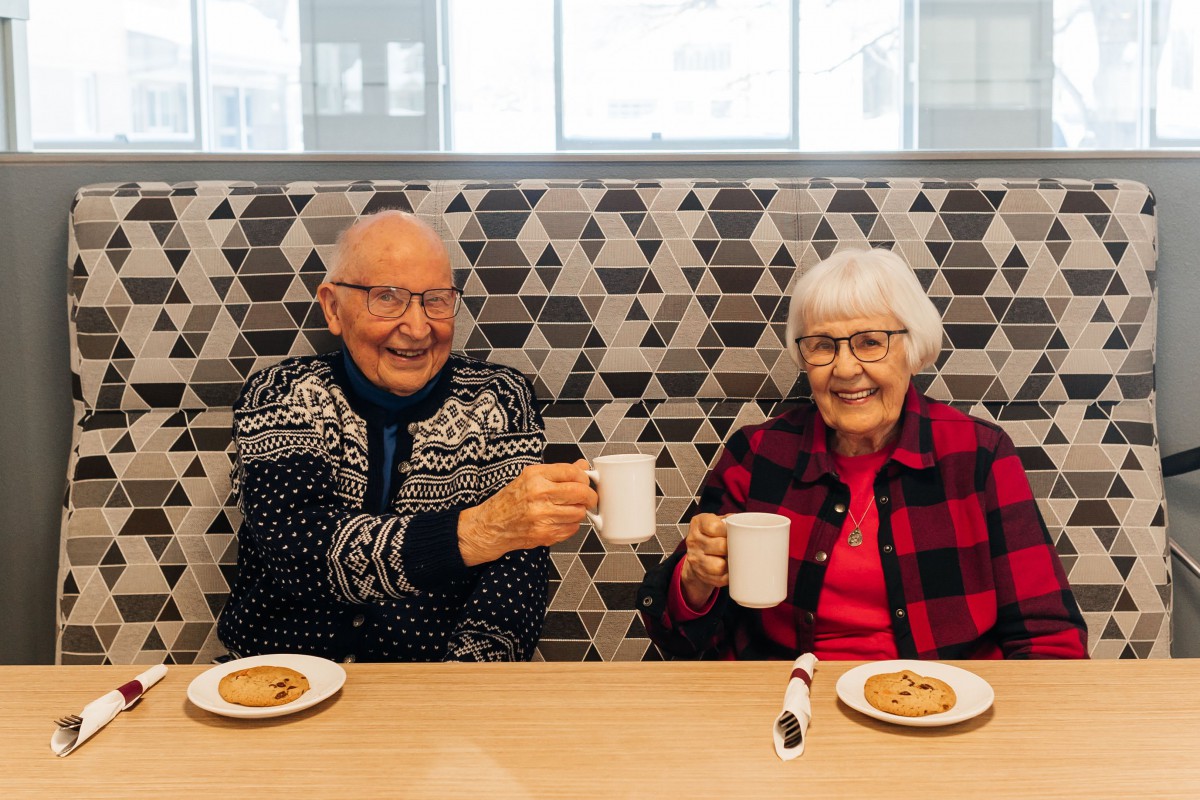 An elderly couple enjoys coffee and cookies, raising their mugs in a cheerful toast.