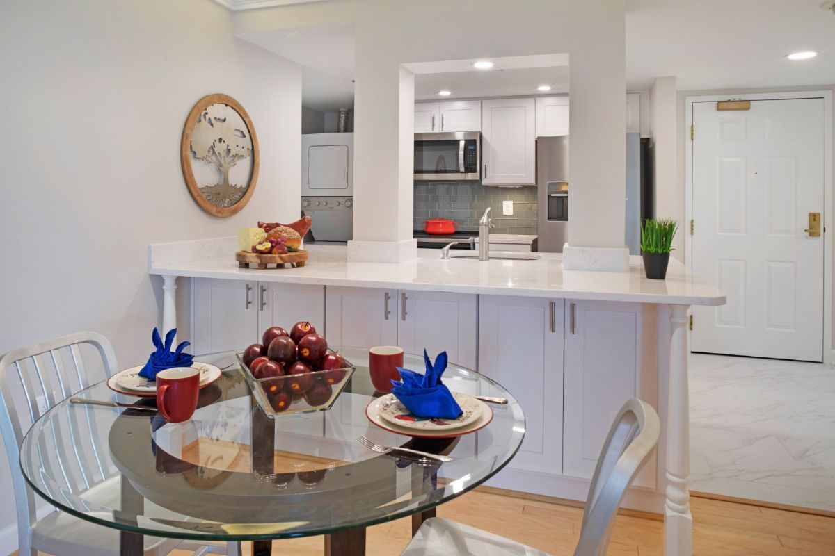 A modern kitchen with an open dining area, featuring a glass table set with blue and red dishware and a bowl of apples.