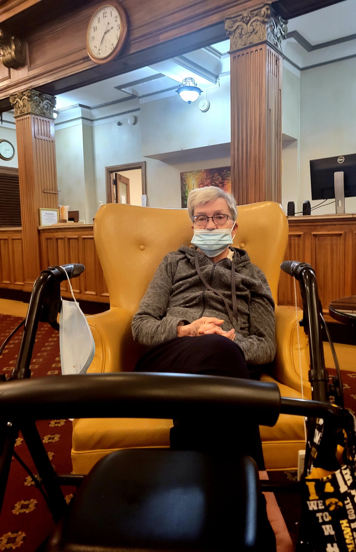 An elderly person wearing a mask sits comfortably in a living room area with wooden paneling.
