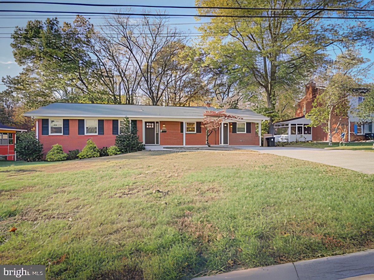 A single-story brick house with a large front lawn and surrounding trees in a suburban neighborhood.
