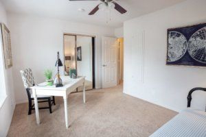 A cozy bedroom with a desk and mirror, featuring a ceiling fan and carpeted floor.