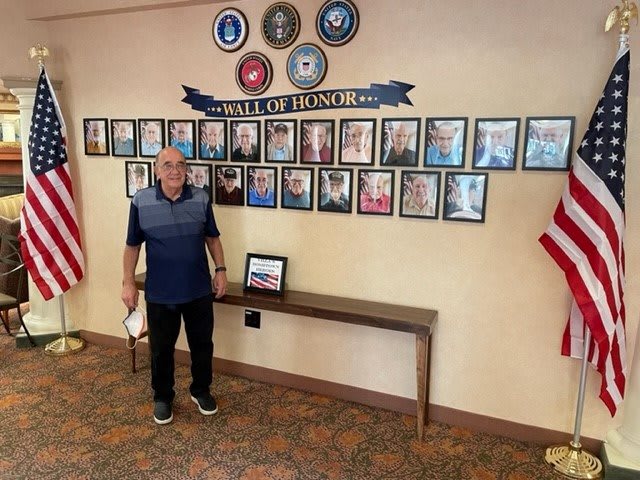 A man stands beside a Wall of Honor displaying photos of veterans and military symbols.