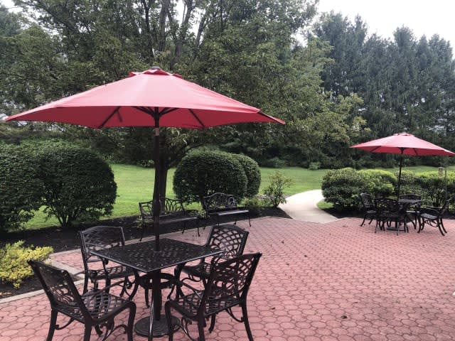 A peaceful patio area with tables, chairs, and red umbrellas set against a lush garden backdrop.