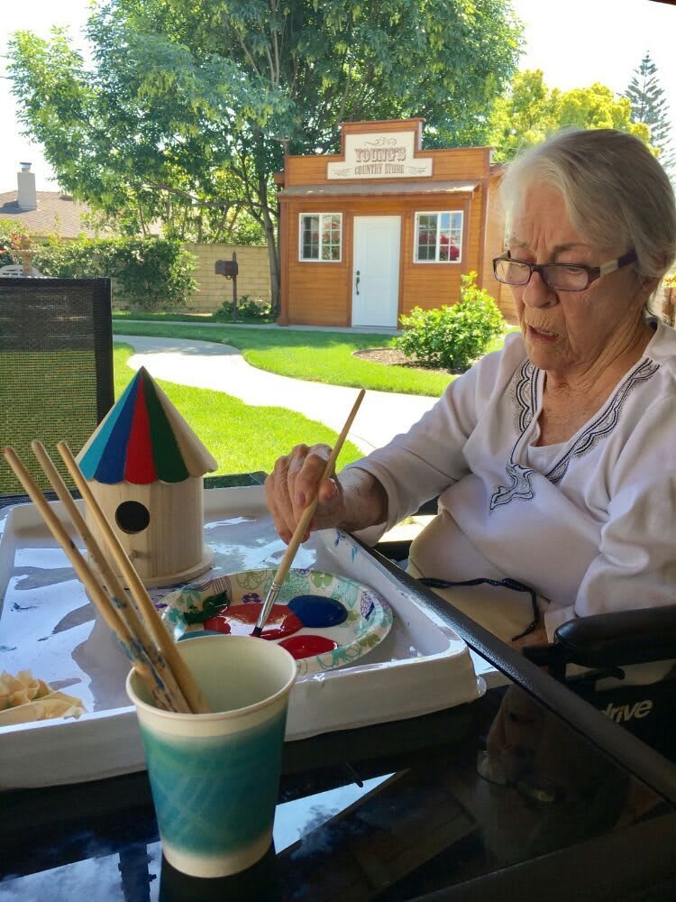 An elderly woman is painting outdoors with various art supplies on a sunny day.