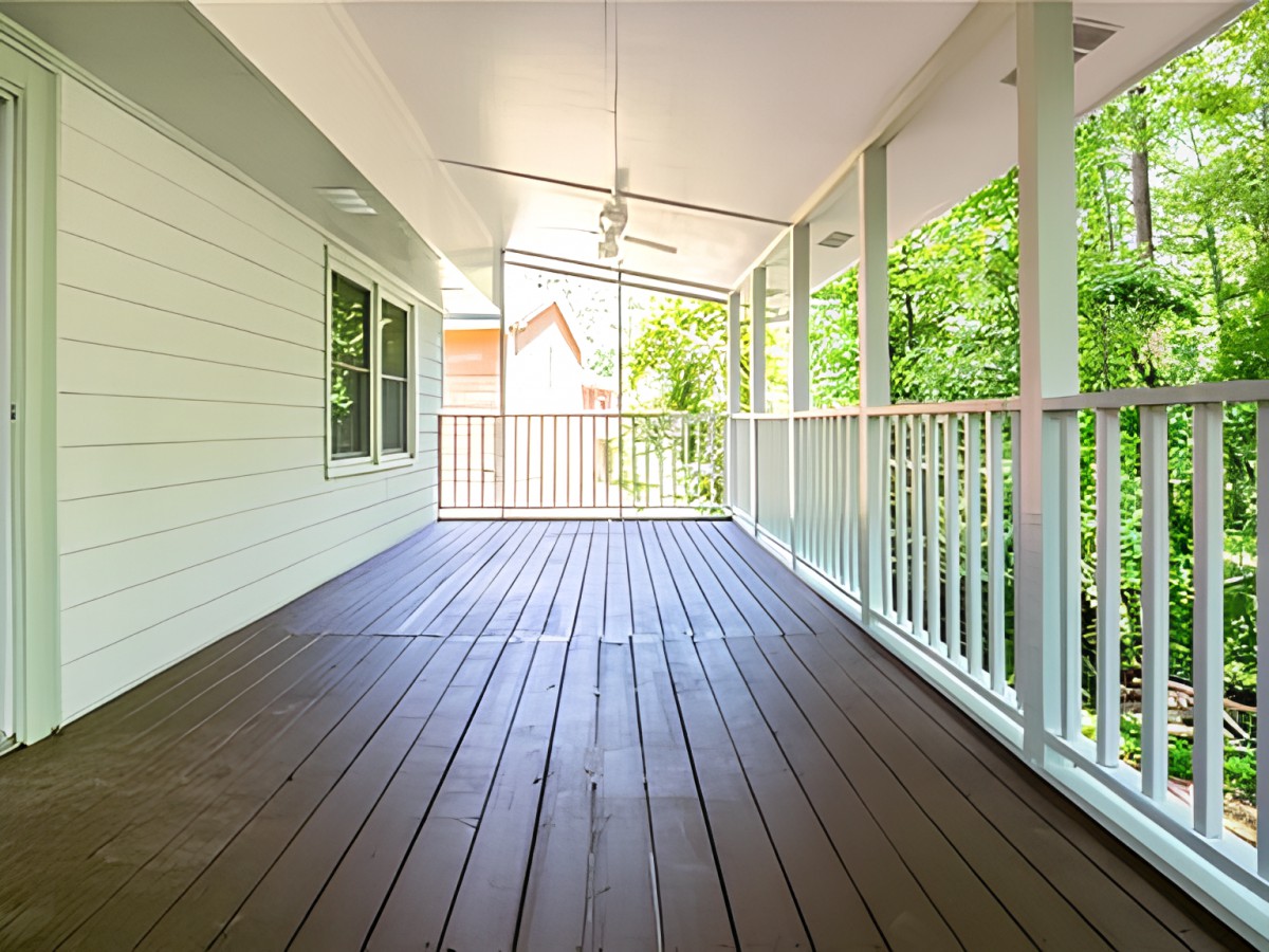 A spacious covered porch with a wooden floor and white railing, surrounded by greenery.