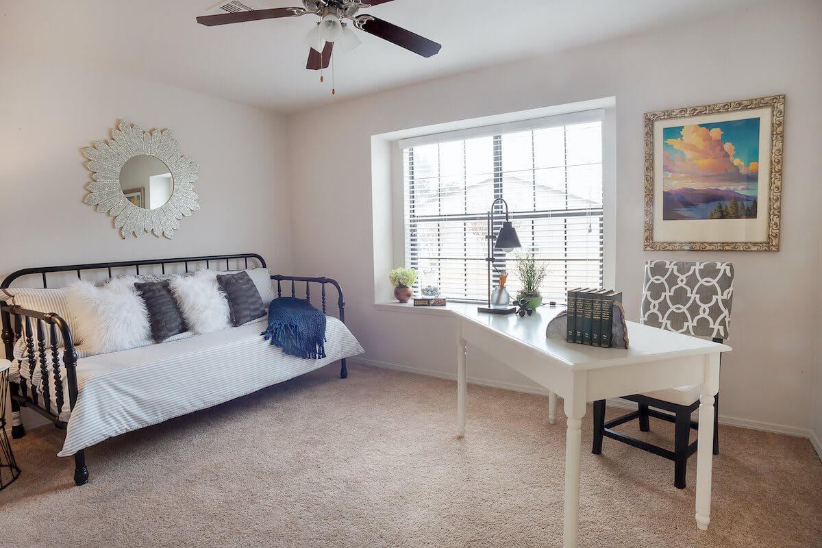 A cozy bedroom with a daybed, a white desk beside a large window, decorated with indoor plants and neutral-colored decor.