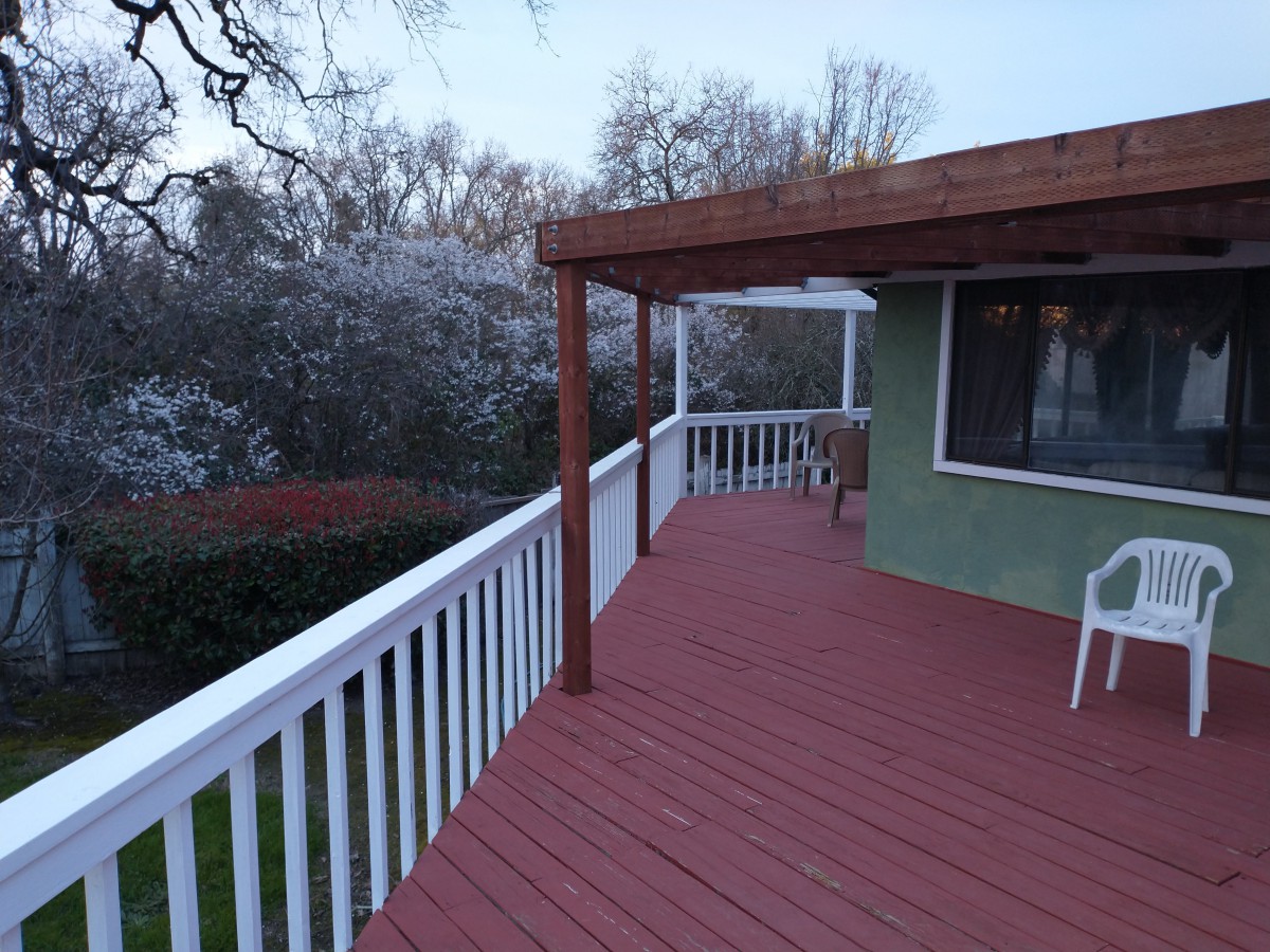 An outdoor wooden deck with white railings and a pergola, surrounded by trees.