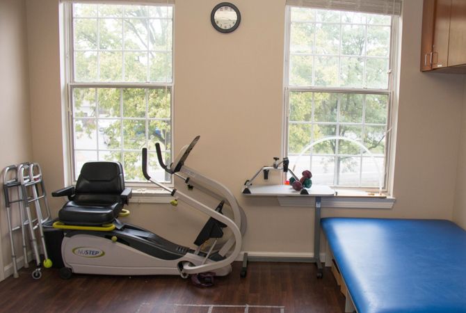 A therapy room with an exercise bike, a massage table, and large windows letting in natural light.