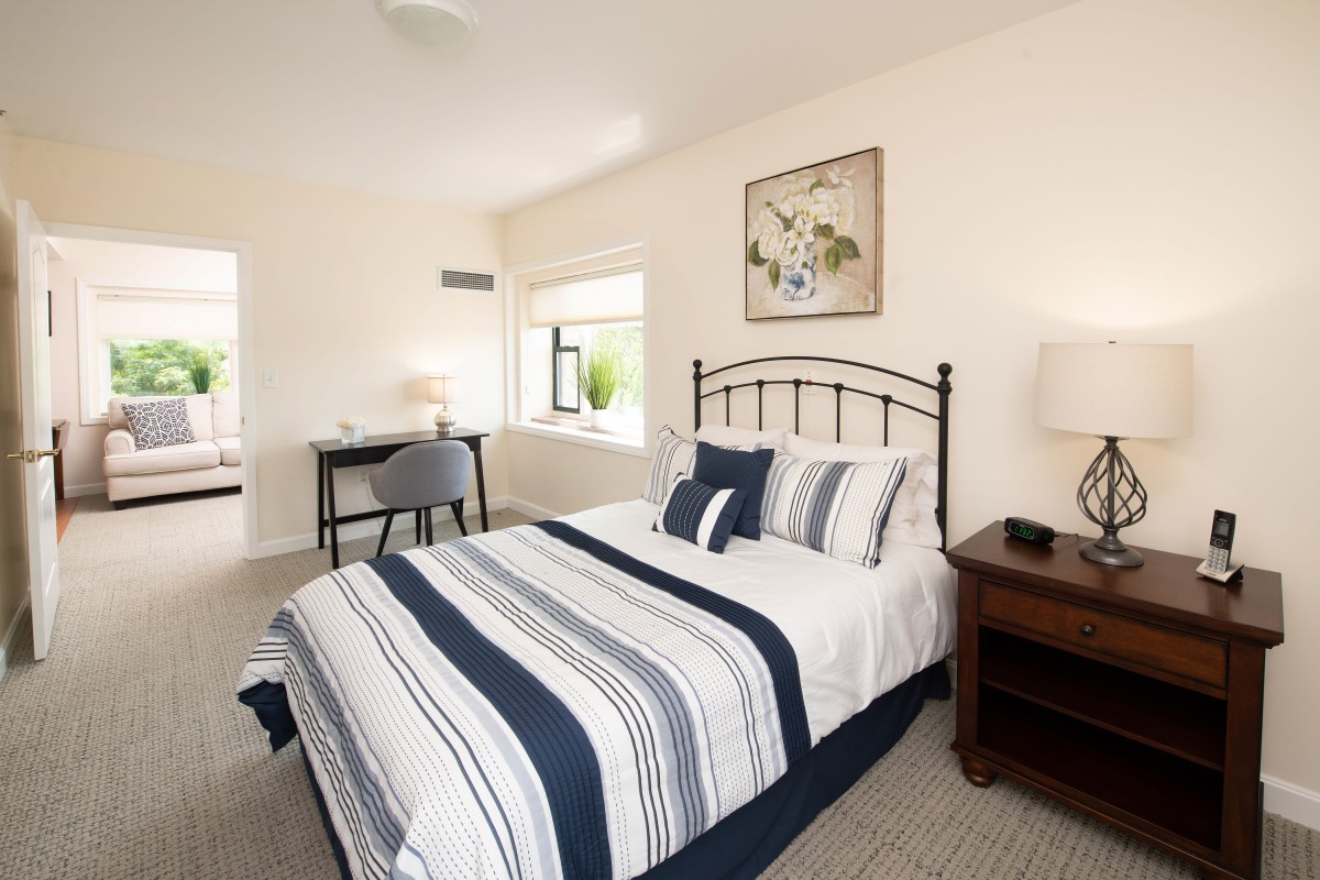 A cozy bedroom with a striped bedspread, a nightstand with a lamp, and a desk near the window.