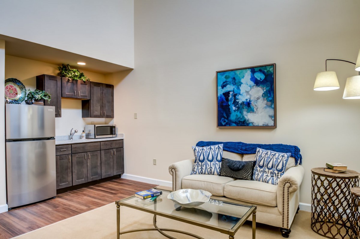 A modern living room with a cozy sofa, vibrant artwork, and a small kitchenette.