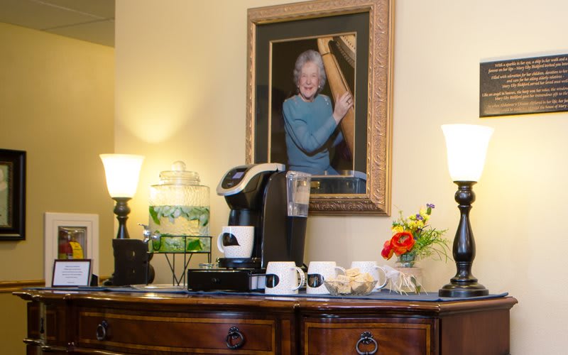 A cozy coffee station setup with a portrait, lamps, and fresh flowers on a wooden dresser.