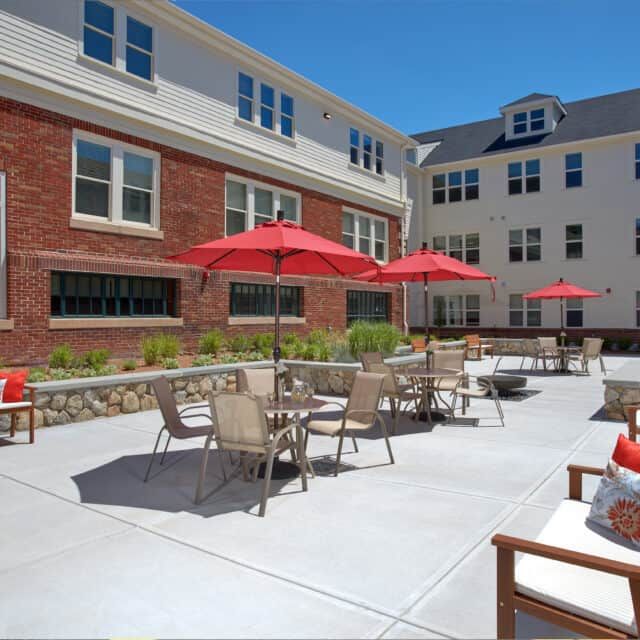 A sunny outdoor patio with tables, chairs, and red umbrellas in front of a brick building.