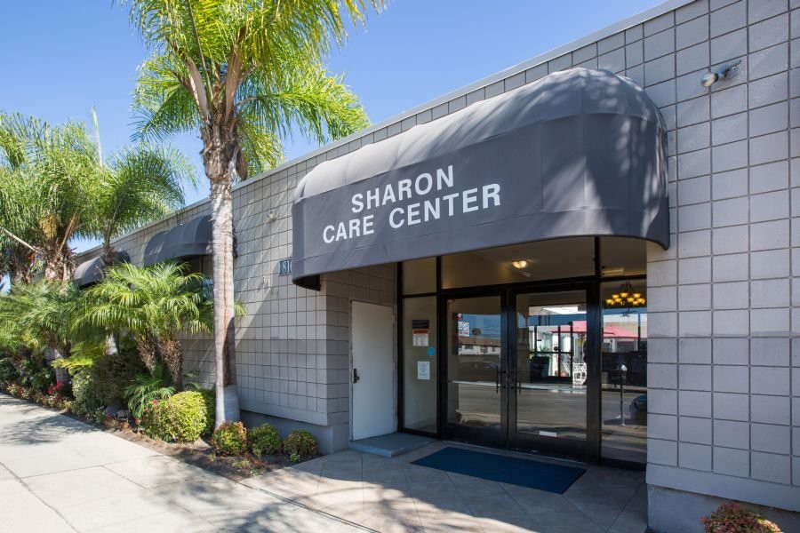 Entrance of Sharon Care Center with palm trees and a sunny sky.