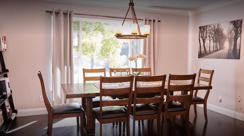 A cozy dining room featuring a wooden table and chairs, illuminated by a chandelier with natural light coming through large windows.