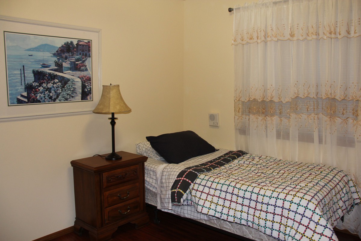 A cozy bedroom with a single bed and a nightstand featuring a lamp, next to a window with sheer curtains.