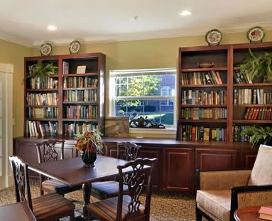 A cozy library room with bookshelves, a table, and chairs, warmly lit by natural light through the window.