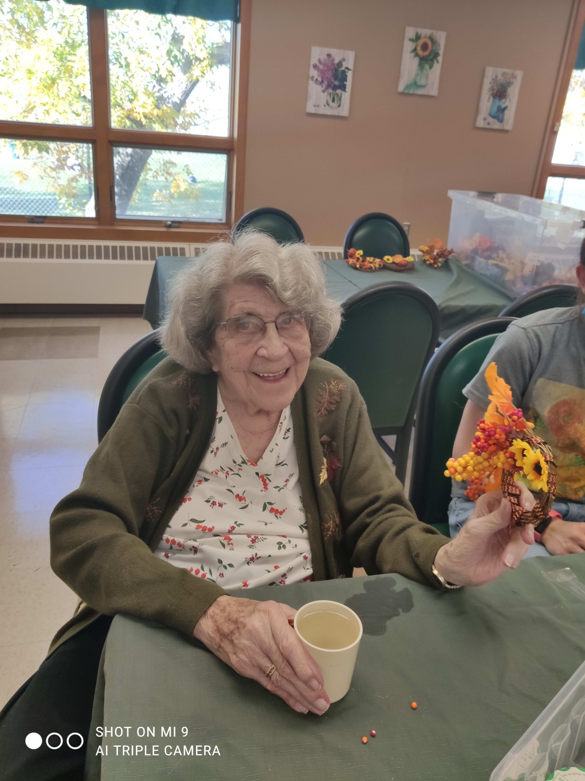 An elderly woman sitting at a table, smiling and holding an autumn-themed decoration indoors.