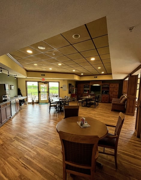 A spacious and cozy living room with wood flooring, featuring a kitchen area, seating, and a large window offering natural light.