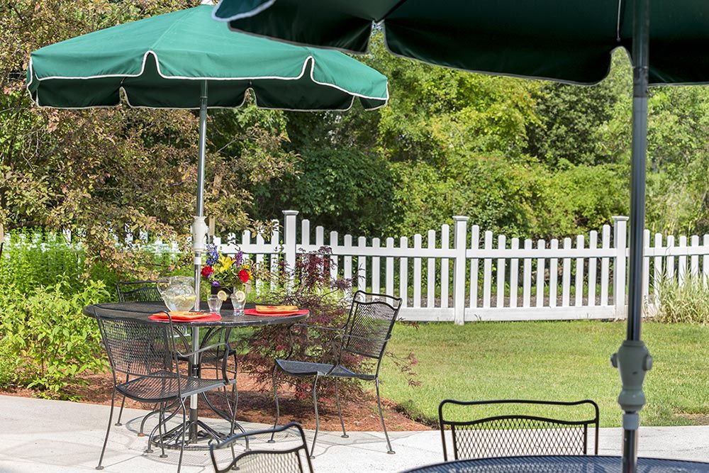 A tranquil outdoor patio with tables, chairs, and umbrellas, surrounded by a white picket fence and lush greenery.