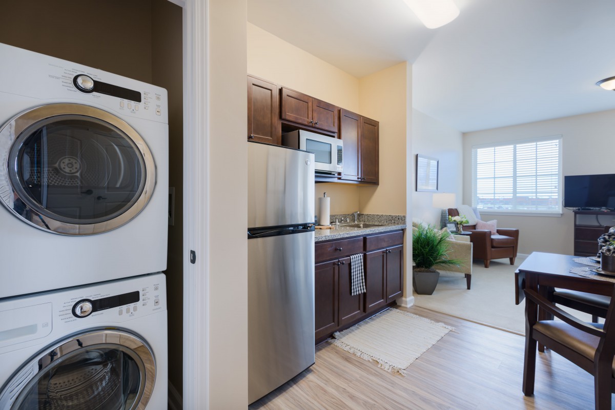 A modern apartment kitchen with stainless steel appliances and a stacked washer and dryer, leading into a cozy living room.