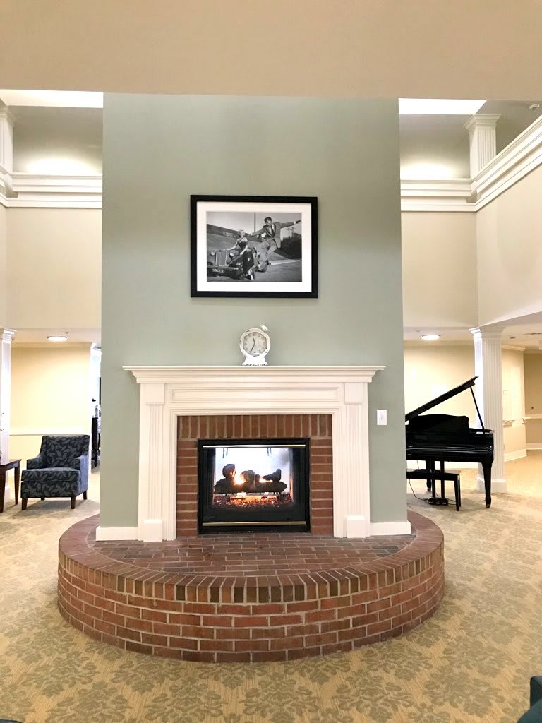 A cozy interior featuring a brick fireplace with a painting above, surrounded by seating and a grand piano.