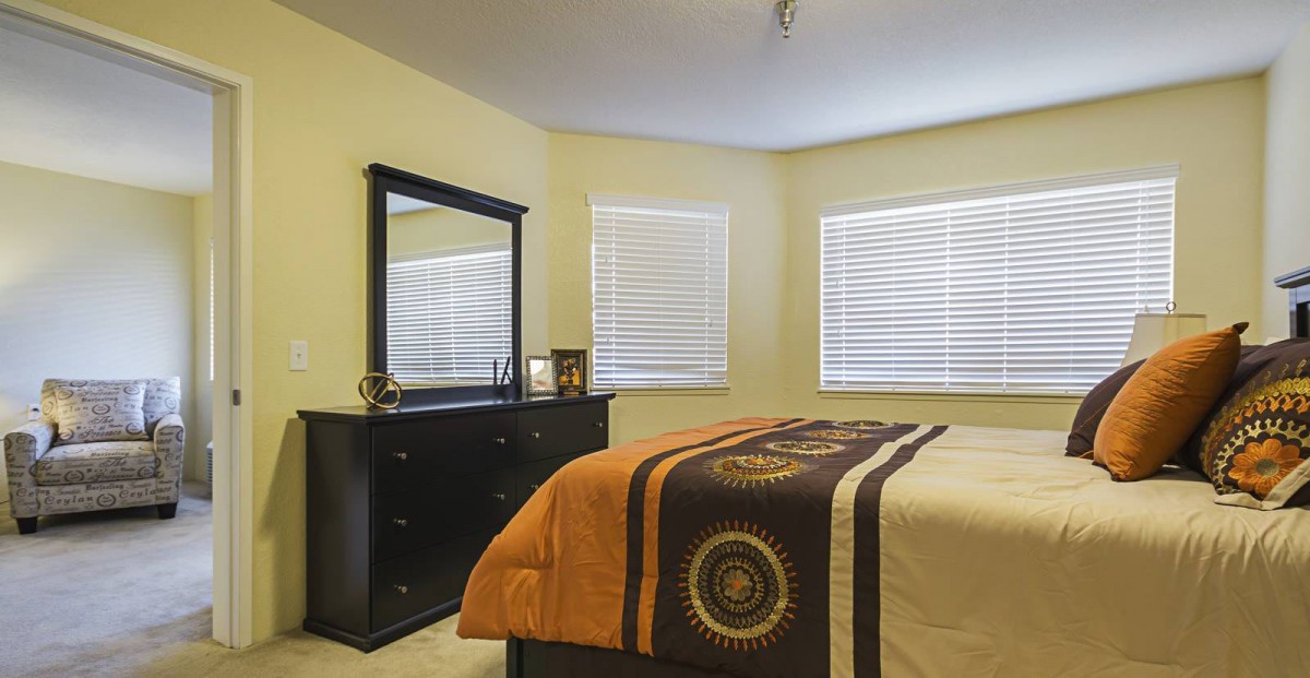 A cozy bedroom with a dresser and mirror, featuring large windows with blinds.
