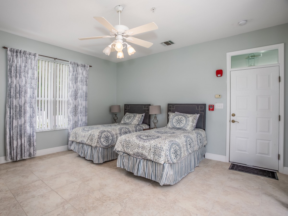 A tidy bedroom with twin beds and a ceiling fan, featuring tiled floors and a window with curtains.
