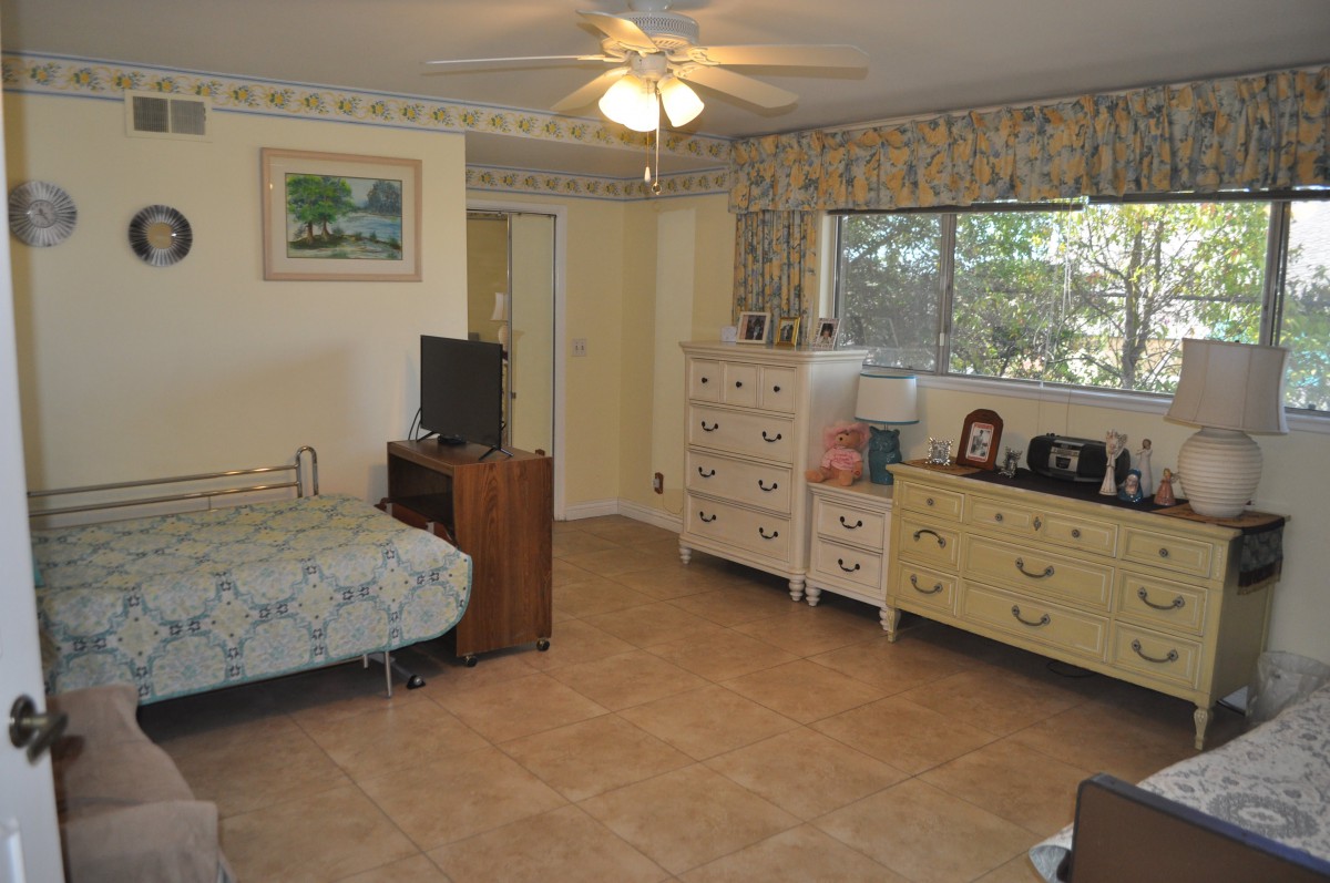 A cozy bedroom with a bed, dresser, and ceiling fan, featuring a large window and decorative elements.