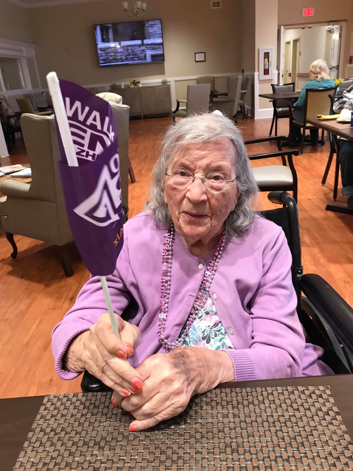 An elderly woman seated indoors, holding a purple flag and smiling.