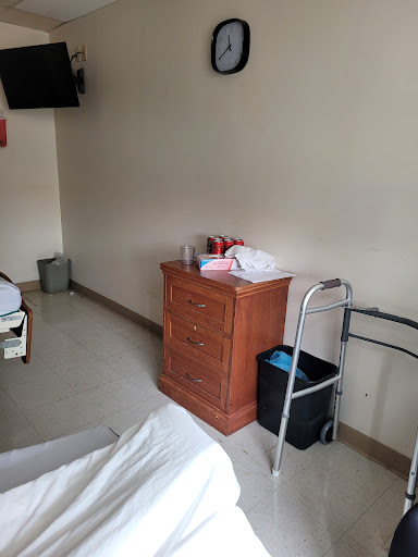 A simple hospital room featuring a bed, a nightstand, a walker, and a trash can.