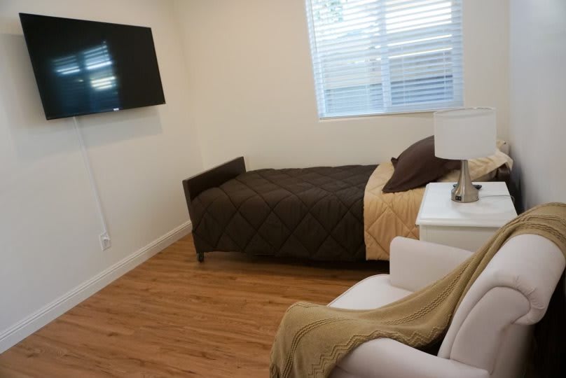 A cozy bedroom with a single bed, wall-mounted TV, and natural lighting from the window.