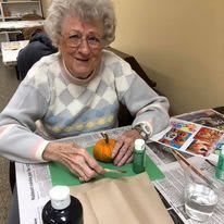 An elderly woman enjoys painting a small pumpkin as part of an indoor arts and crafts activity.