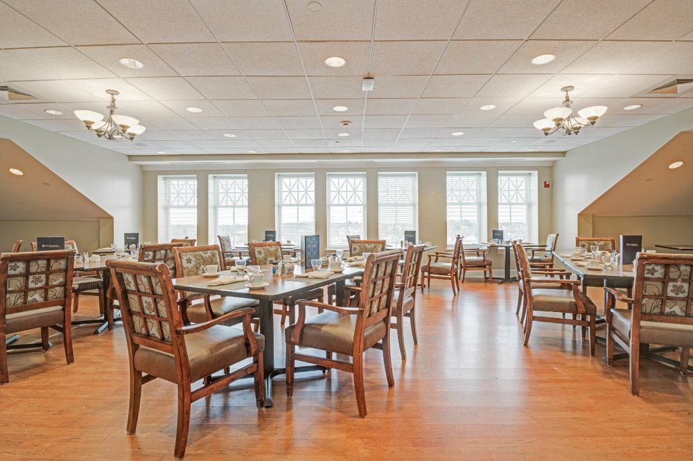 A spacious and well-lit dining room with neatly arranged tables and chairs, featuring large windows and wooden flooring.