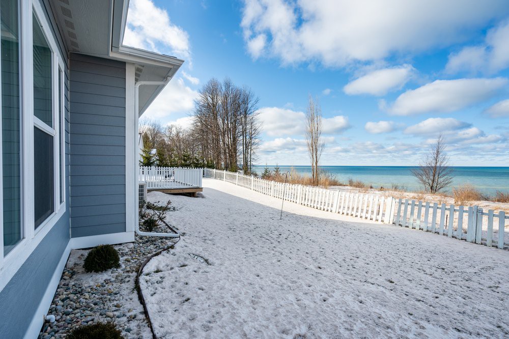 A snowy beach view with a house and fence near a lake under a partly cloudy sky.