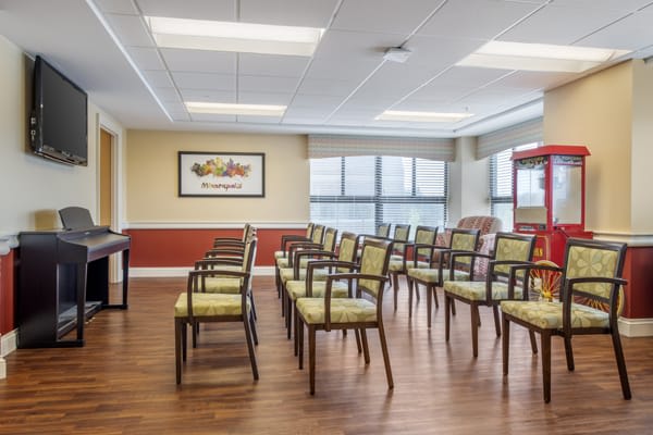 An empty lecture or meeting room with rows of chairs facing the front, featuring large windows and a wooden floor.
