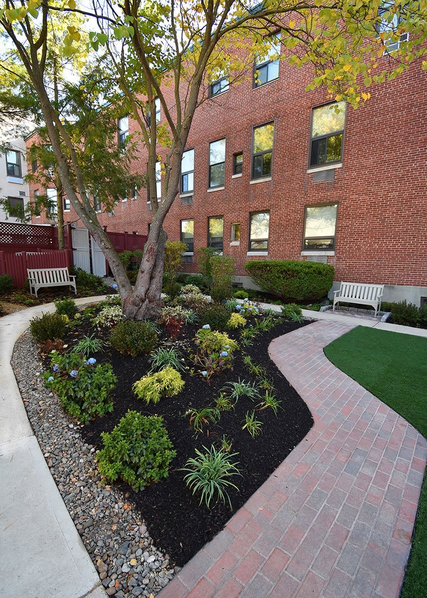 A landscaped garden with pathways, benches, and a variety of plants is situated next to a brick building.