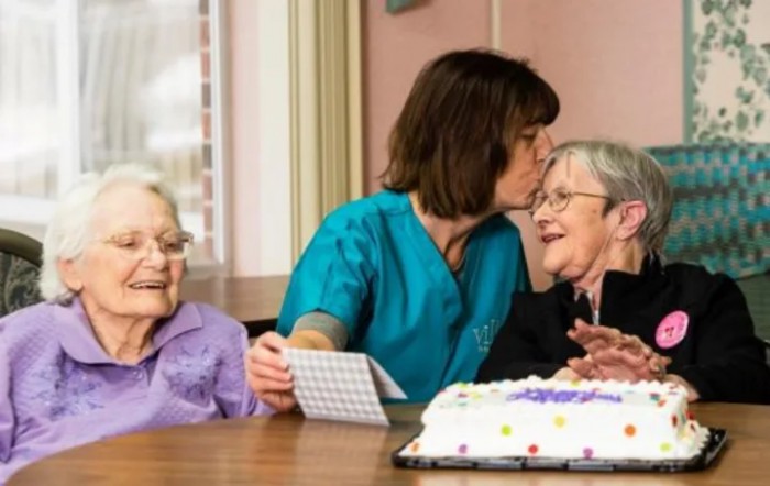 A caregiver shares a tender moment with elderly women at a celebration, accompanied by a cake on the table.