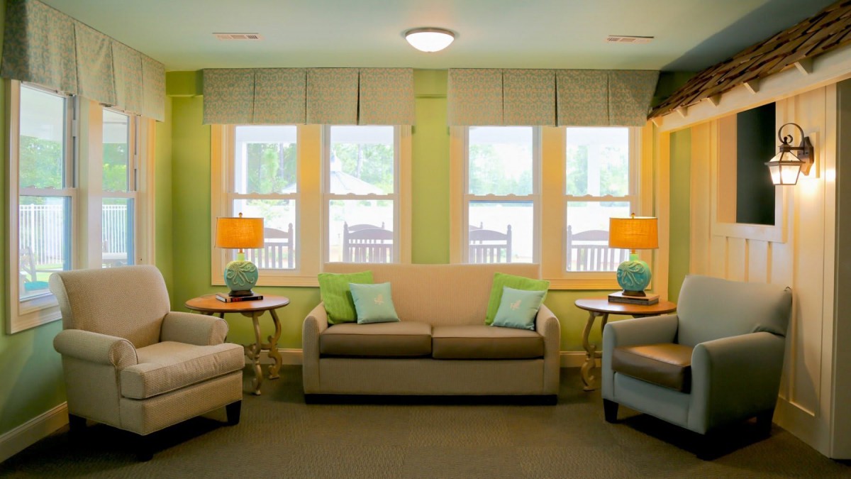 A cozy living room with sofas and armchairs arranged around a set of windows, complemented by warm lighting from table lamps.