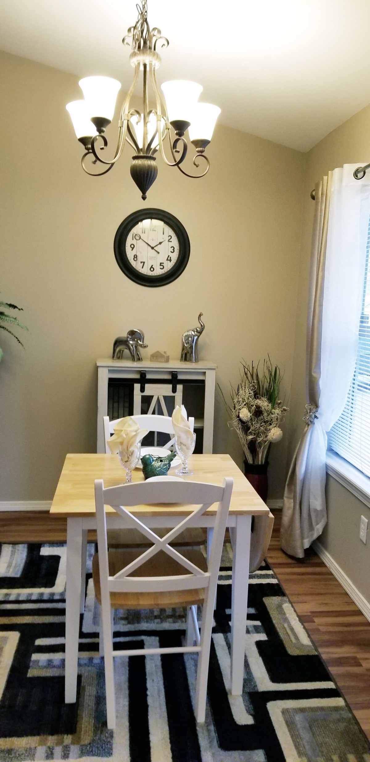 A cozy dining area with a small table and chairs, adorned with decorative items and plants, featuring a wall clock.